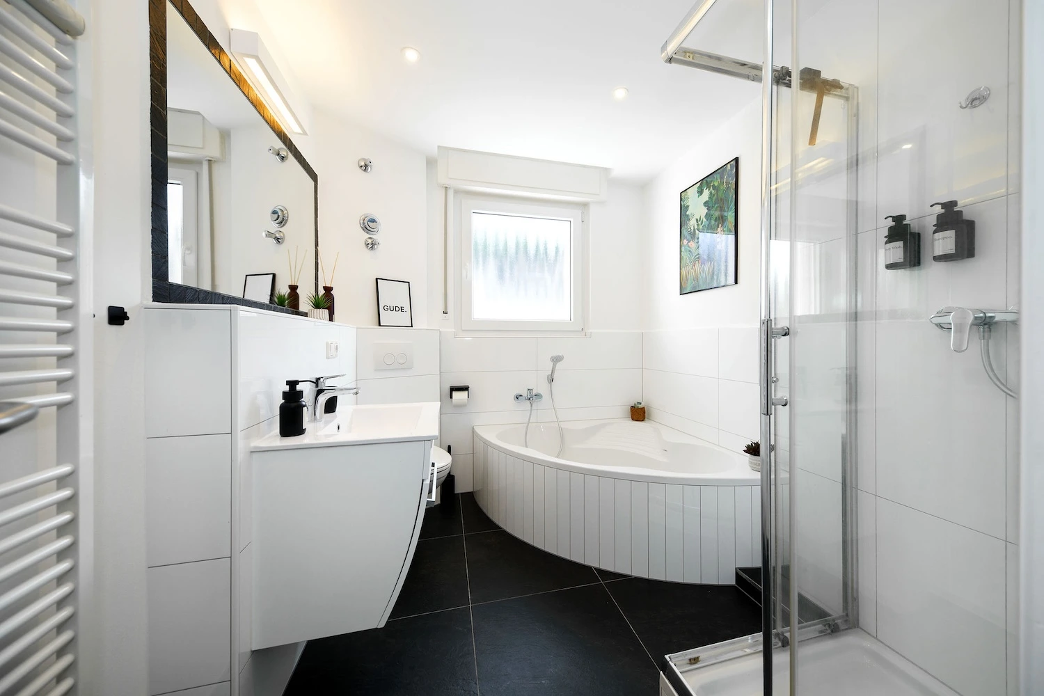 Modernes Badezimmer mit weißer Eckbadewanne, separater Glasdusche, Waschbecken und schwarzem Fliesenboden.