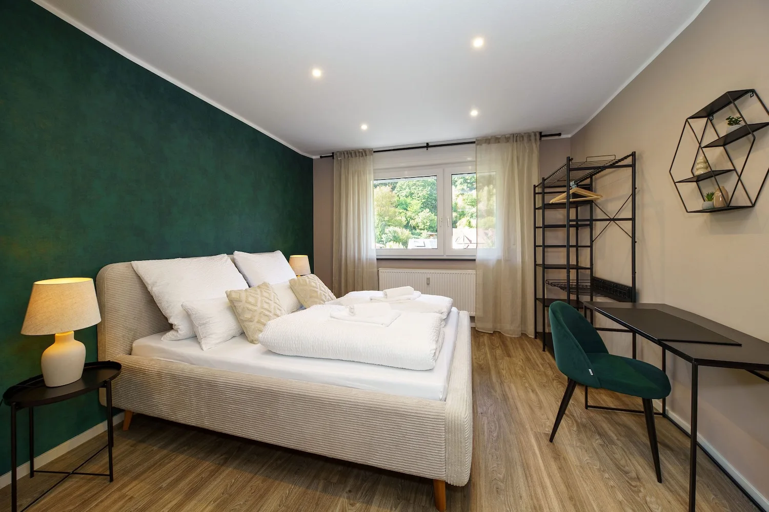 Modernes Schlafzimmer mit weißem Doppelbett, dunkelgrüner Akzentwand, Schreibtisch, grünem Stuhl und Fenster.