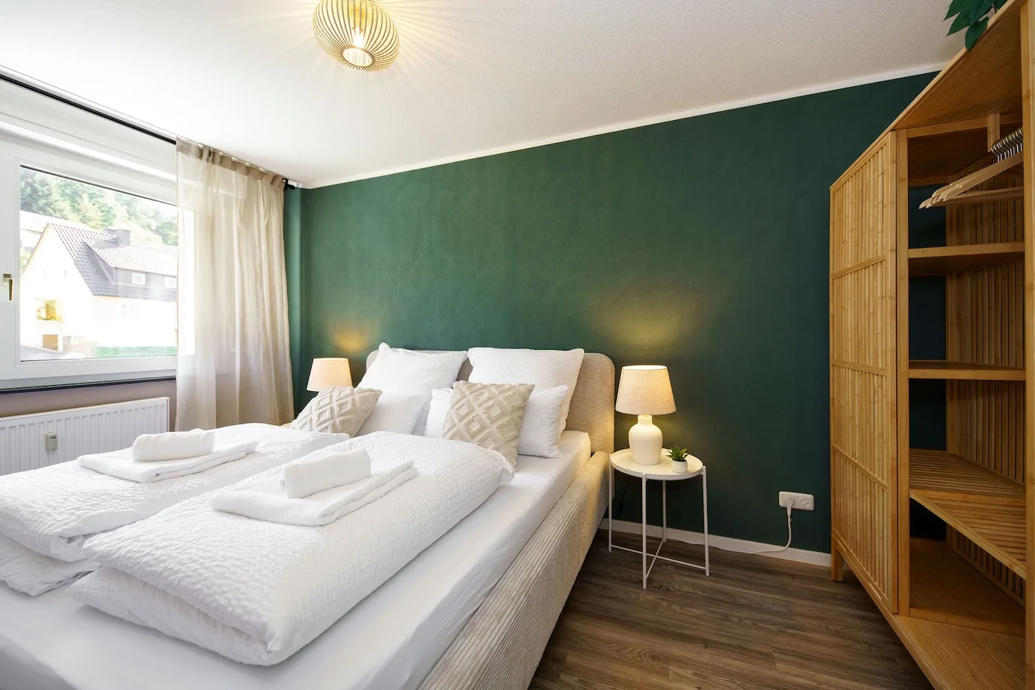 Helles Schlafzimmer mit weiß bezogenem Doppelbett, dunkelgrüner Wand, Nachttisch und offenem Holzkleiderschrank.