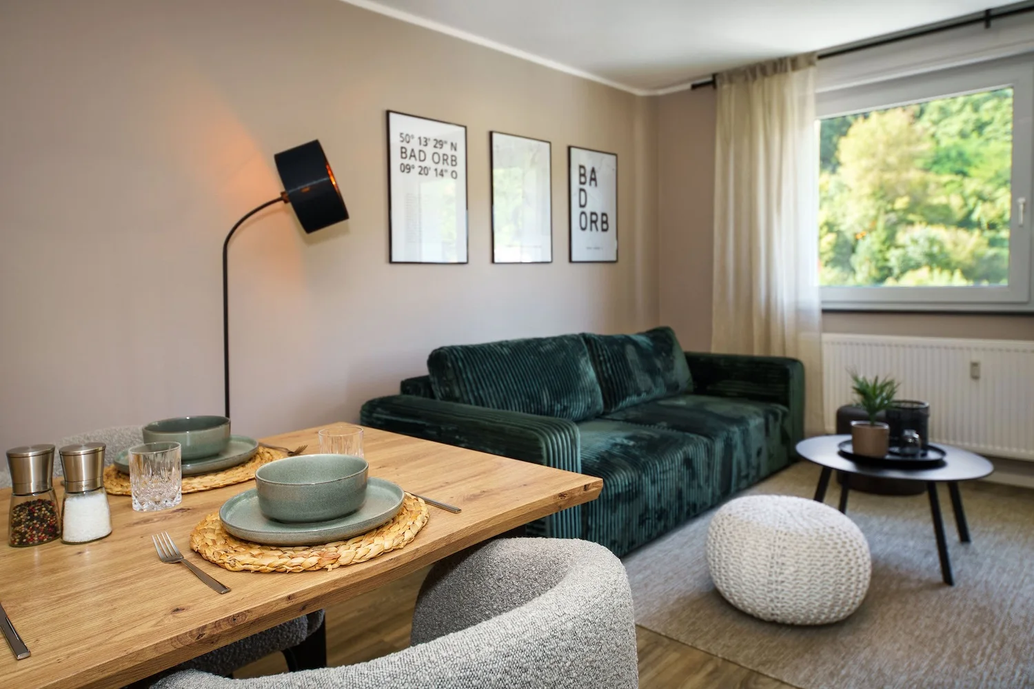 Gemütliches Wohnzimmer mit dunkelgrünem Sofa, gedecktem Esstisch, Stehlampe und Fenster mit Grünblick.